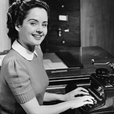 A female secretary smiles briskly at the camera as she sits typing at  a desk in a 1950s office. 