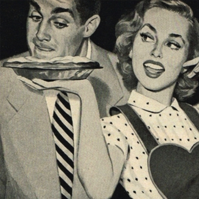 In a black and white pastiche of a 1950s commercial, a pretty housewife in an apron offers up a freshly cooked pie to her husband, who leans forward to  smell it approvingly. 